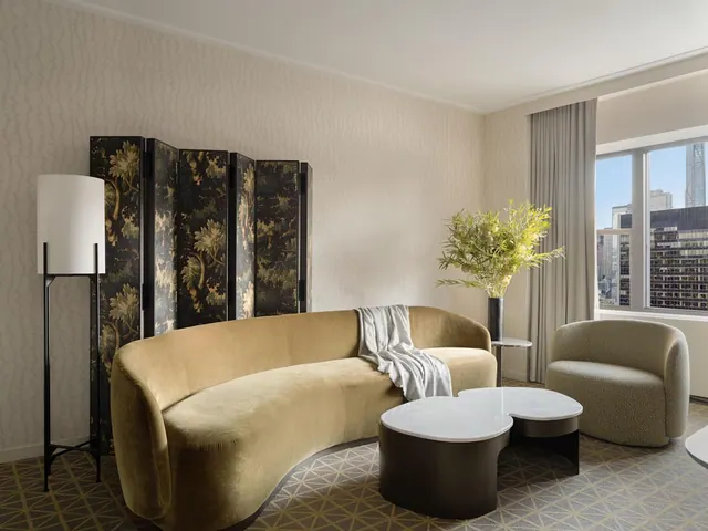 A living area with a curved gold sofa, a folding screen, a floor lamp, two low coffee tables and an armchair, with a window showing city buildings.