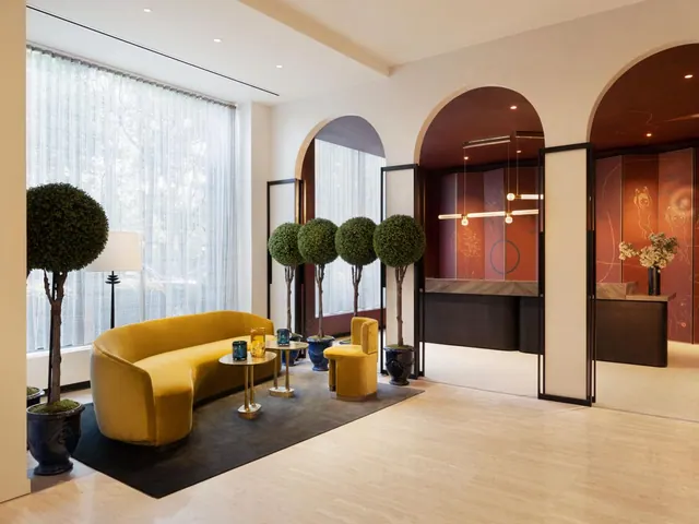 A lobby seating area with a curved yellow velvet sofa, low tables and topiary trees, with a reception desk visible through an arched doorway.