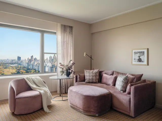 A living area with a pink velvet sectional sofa, armchair and ottoman, and a large window looking over Central Park and the city skyline.