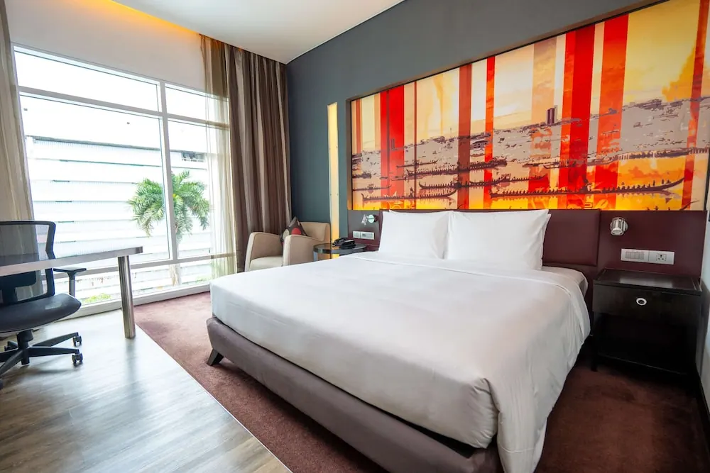 A bedroom with a king bed under a large colourful boat artwork, a work desk and armchair by a floor-to-ceiling window, and a palm tree and building wall visible outside.