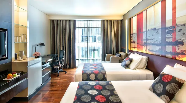 A hotel room with two beds with dotted runners, a desk and chair, an armchair, a wall-mounted television and a large boat artwork above the beds.