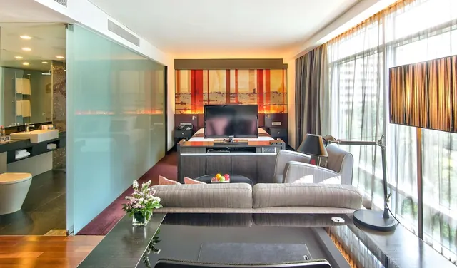 A hotel suite living area with a grey sofa, armchair, wall-mounted television, and a glass-partitioned bathroom with a basin and toilet on the left.