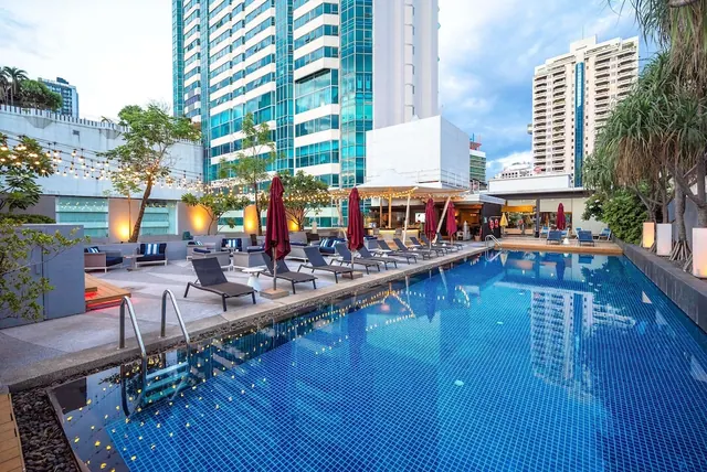 Outdoor swimming pool with lounge chairs, closed red parasol umbrellas and string lights, with high-rise towers behind at dusk.