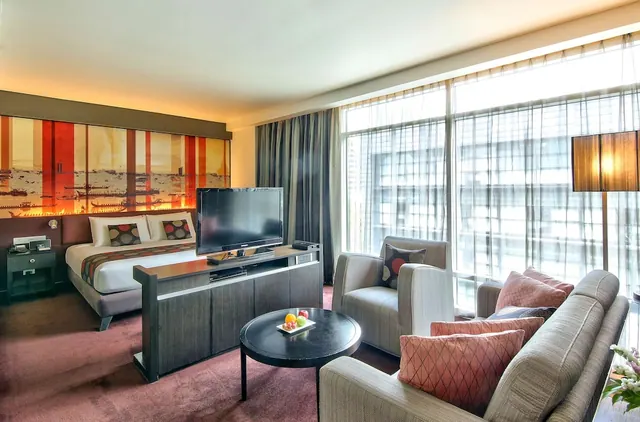 A hotel room with a king bed, a flat-screen TV on a wooden cabinet, a grey sofa and armchairs, a round coffee table with a fruit plate, and floor-to-ceiling windows with sheer curtains looking onto nearby buildings.