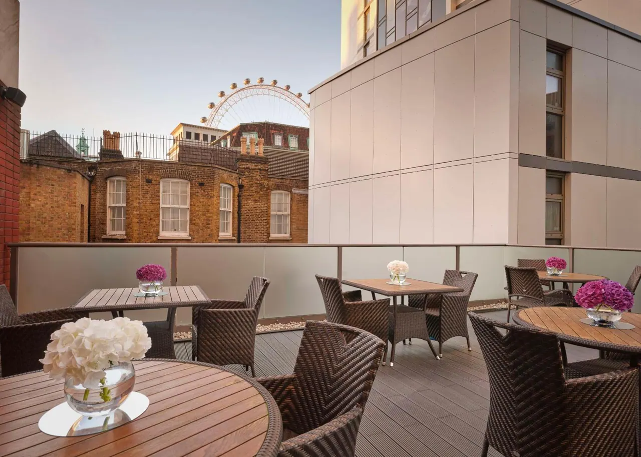 A rooftop terrace with wicker chairs and wooden tables holding flower vases, with the London Eye visible above brick buildings.