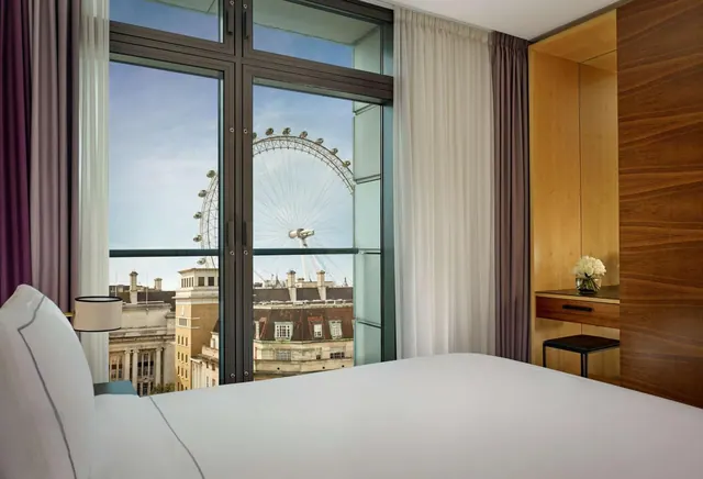 A bedroom with a made bed facing floor-to-ceiling windows that frame the London Eye and nearby rooftops.