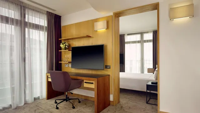 A hotel room with a wall-mounted television above a wooden desk and purple chair, with a bed visible through a doorway to the right.