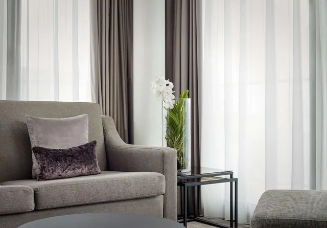 A grey upholstered armchair with cushions sits beside a side table holding a white orchid in a glass vase, with sheer and taupe curtains behind.