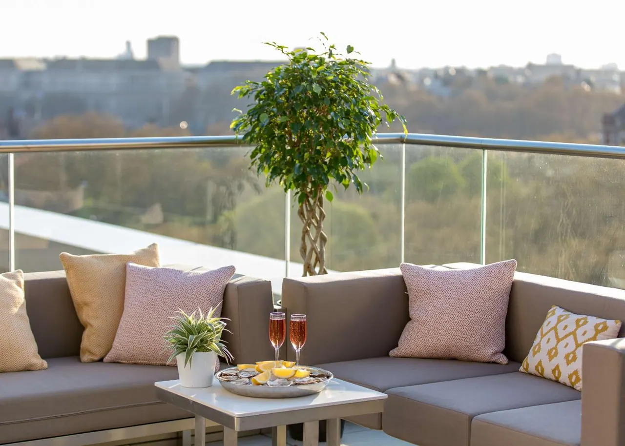 A rooftop terrace with a grey sectional sofa, patterned cushions, a potted tree, and a side table holding champagne flutes and a plate of oysters, with a glass balustrade and a hazy city view behind.