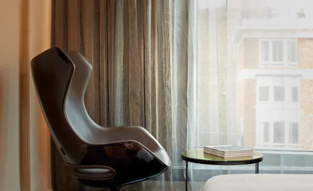 A dark leather armchair stands by a window with sheer and linen curtains, beside a small round side table holding a stack of books, with a neighbouring building facade visible outside.