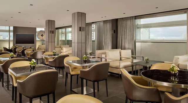 A lounge area with leather armchairs, marble-topped tables, cream sofas and a wall-mounted television, with city buildings visible through the windows.
