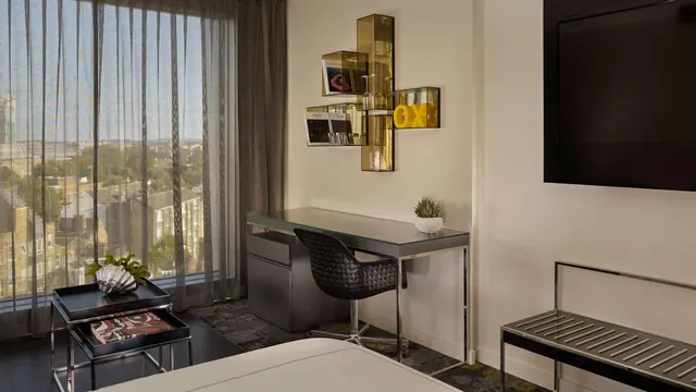 A hotel bedroom with a desk and chair, a wall-mounted television, and a window overlooking residential rooftops and trees.