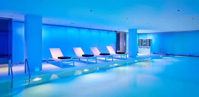 An indoor pool lit in blue, with a row of white sun loungers with navy cushions along its edge.
