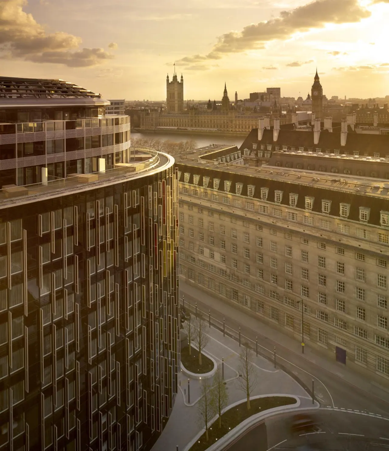Elevated view at sunset over a curved modern building and older stone buildings toward the Palace of Westminster and Big Ben, with the River Thames beyond.