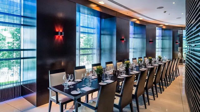 A long dining table set with glasses, plates and napkins in a private dining room with dark walls, red wall lights and tall windows showing trees outside.