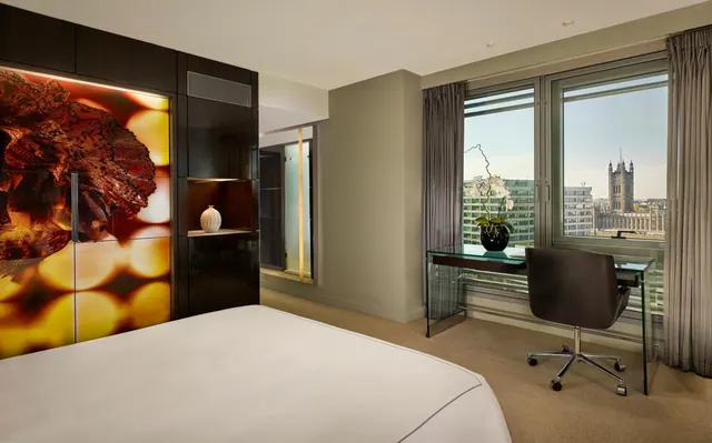 A bedroom with a made bed, an illuminated wall panel, a glass desk and chair, and a window looking out over the Palace of Westminster.