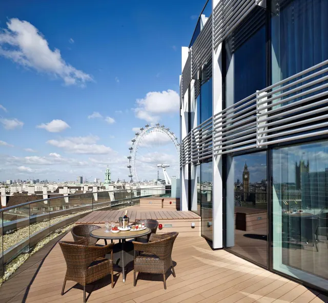 A furnished rooftop terrace with wicker chairs and a round table, with the London Eye and city rooftops visible beyond the glass railing.