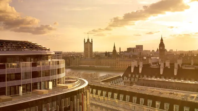 A view over the Thames towards the Palace of Westminster and Big Ben at sunset, with rooftops and a curved building in the foreground.