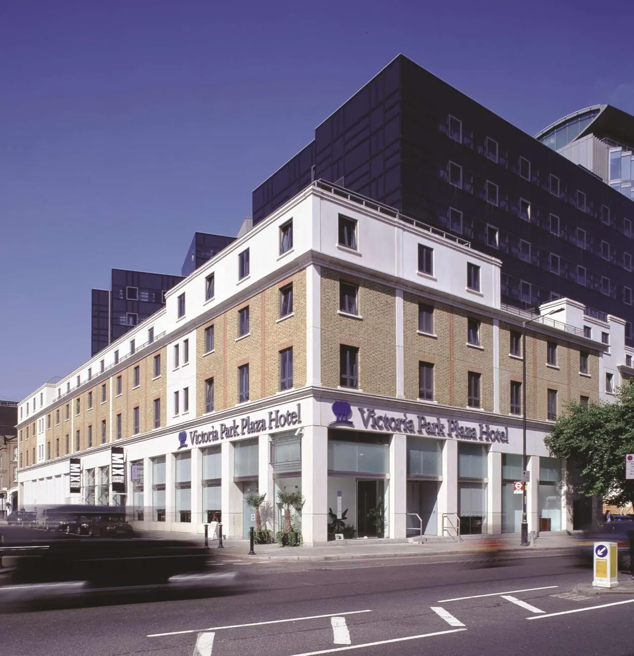 Exterior of the Victoria Park Plaza Hotel, a building with a white and brick lower facade and a dark glass tower behind it, seen from the street under a clear blue sky.