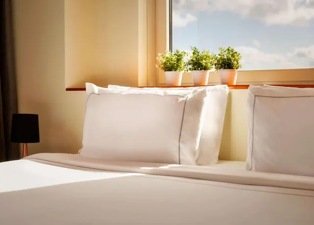 A made bed with white pillows and grey-edged linen sits beside a window with three potted plants on the sill.