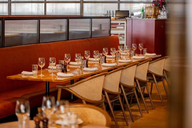 A long dining table set with wine glasses, folded napkins and pepper mills sits beside a tan leather banquette and cane-back chairs in a restaurant.