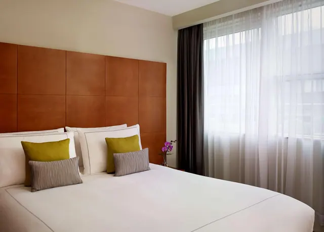 A made-up bed with a wooden panelled headboard, green and grey cushions, and a window with sheer curtains beside it.