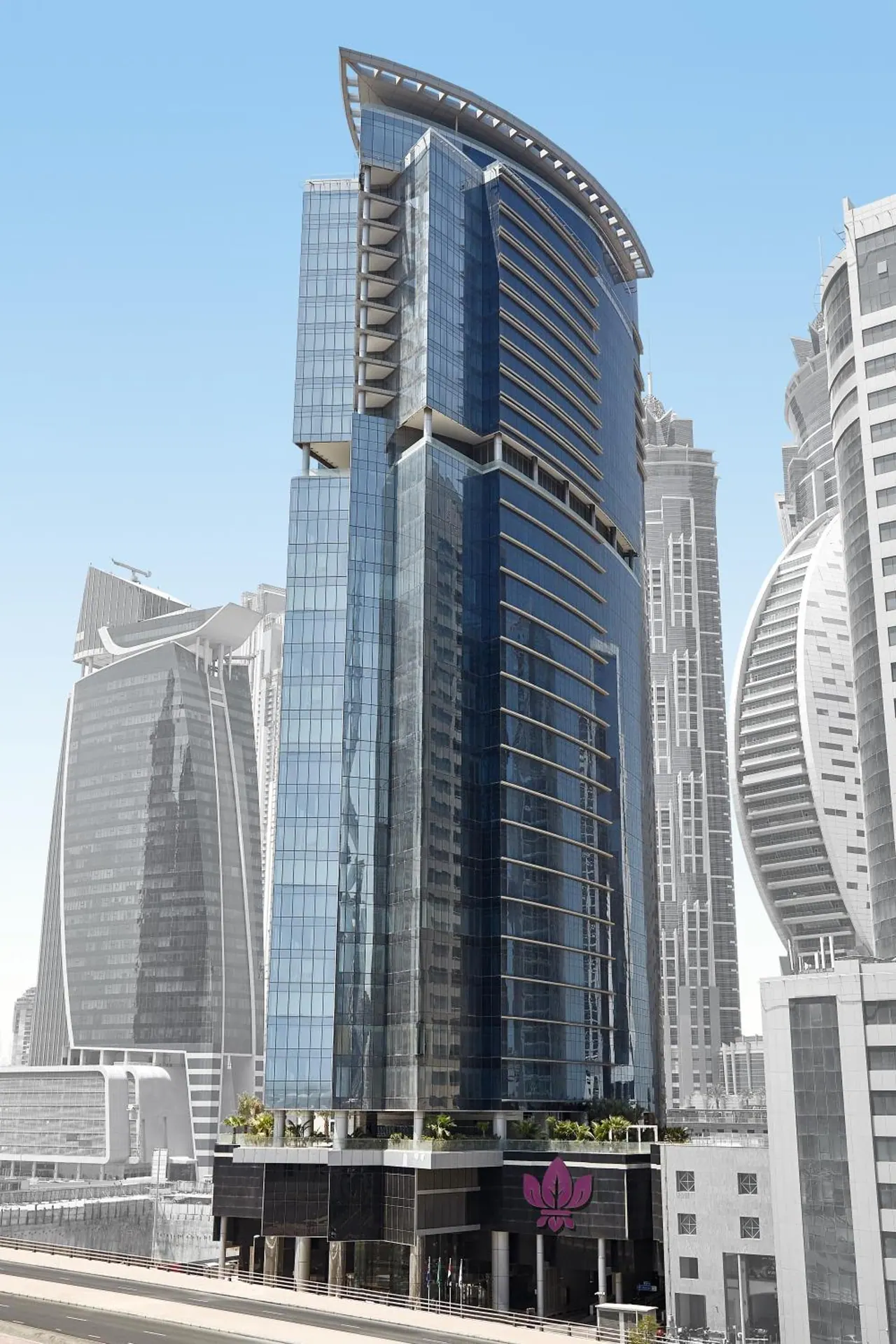 The exterior of a tall curved glass tower with a pink lotus logo at its base, flanked by other high-rise buildings under a clear blue sky.