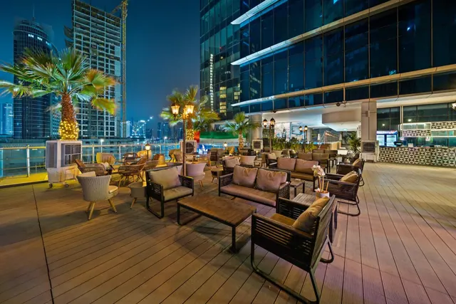 An outdoor lounge terrace at night with sofas, armchairs and coffee tables on a timber deck, lit palm trees and lanterns, a glass tower beside it, and city towers and water visible beyond the railing.