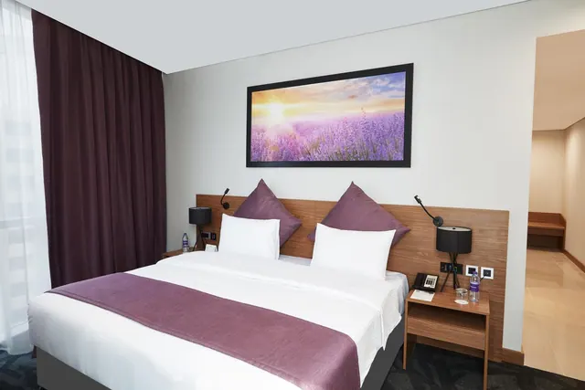 A bedroom with a king bed, a purple runner across the foot, a framed lavender print above the headboard, and bedside lamps and water bottles on the nightstands.