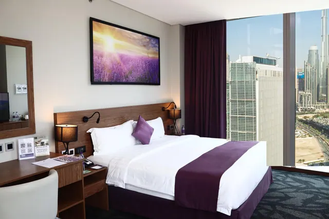 A hotel bedroom with a king bed, a framed artwork on the wall, a desk and mirror, and floor-to-ceiling windows showing high-rise towers outside.