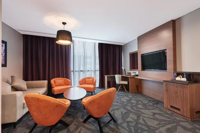 A hotel suite living area with a sofa, orange armchairs around a round table, a wall-mounted television, a desk and a kettle on a wood cabinet.