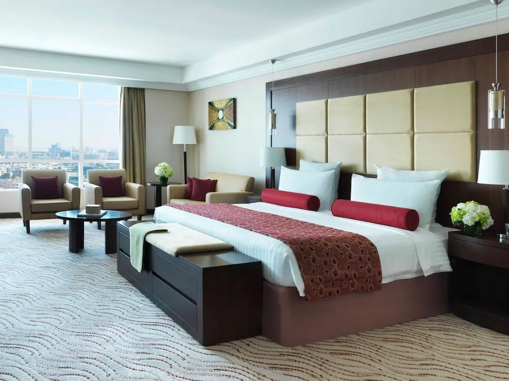 A king bed with red bolsters and a red runner sits beside armchairs and a round coffee table, with floor-to-ceiling windows showing a city skyline.