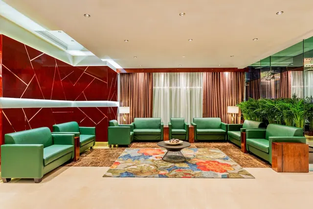 A lobby seating area with green leather sofas and armchairs, a round coffee table with a fruit bowl, and a floral rug in front of a red wall with gold lines.