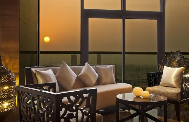 A seating area with a sofa, two carved armchairs, a glass-top side table and candles, with large windows showing a hazy sunset over the city.
