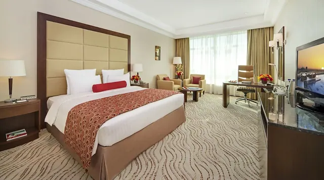 A hotel bedroom with a king bed, two armchairs beside a round coffee table, a desk, and a wall-mounted television.