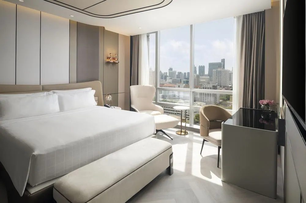 A bedroom with a king bed, armchairs, a desk and a floor-to-ceiling window overlooking a city skyline.