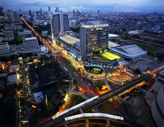 Aerial dusk view over a Bangkok street with the MBK Center building, an elevated Skytrain line and a stadium in the distance.