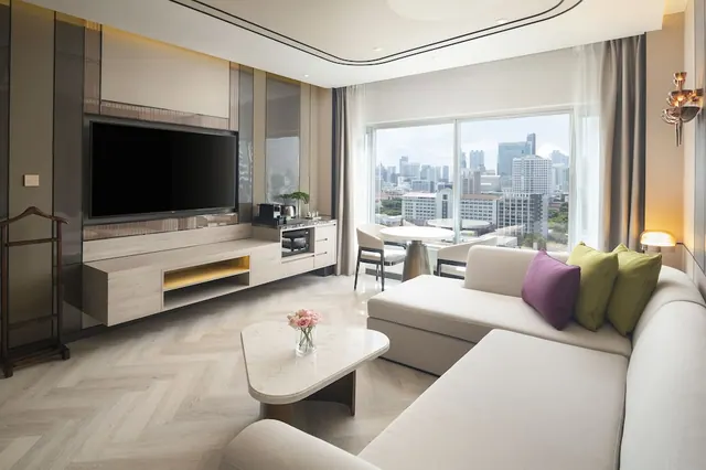 A living area with a wall-mounted television, a cream sofa with cushions, and a large window looking out over a city skyline.