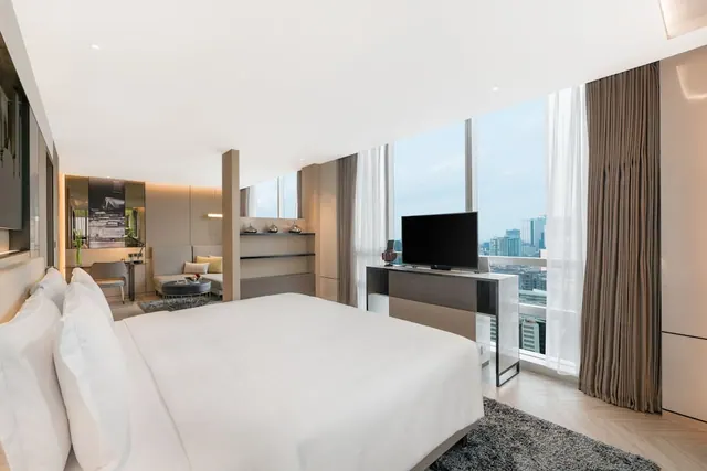 A bedroom with a made king bed, a television on a cabinet beside floor-to-ceiling windows overlooking city high-rises, and a sofa and desk area beyond.