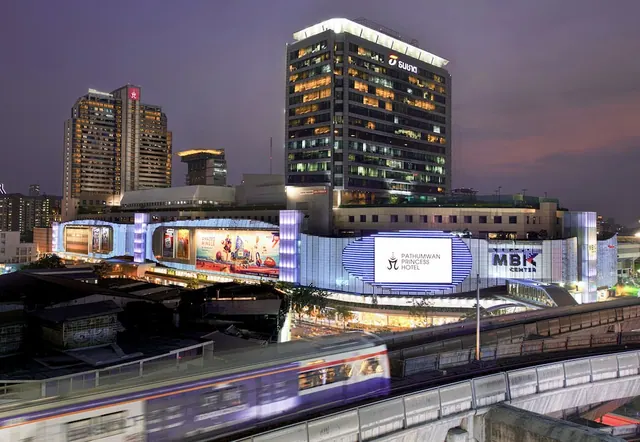 Exterior of the Pathumwan Princess Hotel at dusk, with the MBK Center building alongside and a blurred Skytrain train in the foreground.