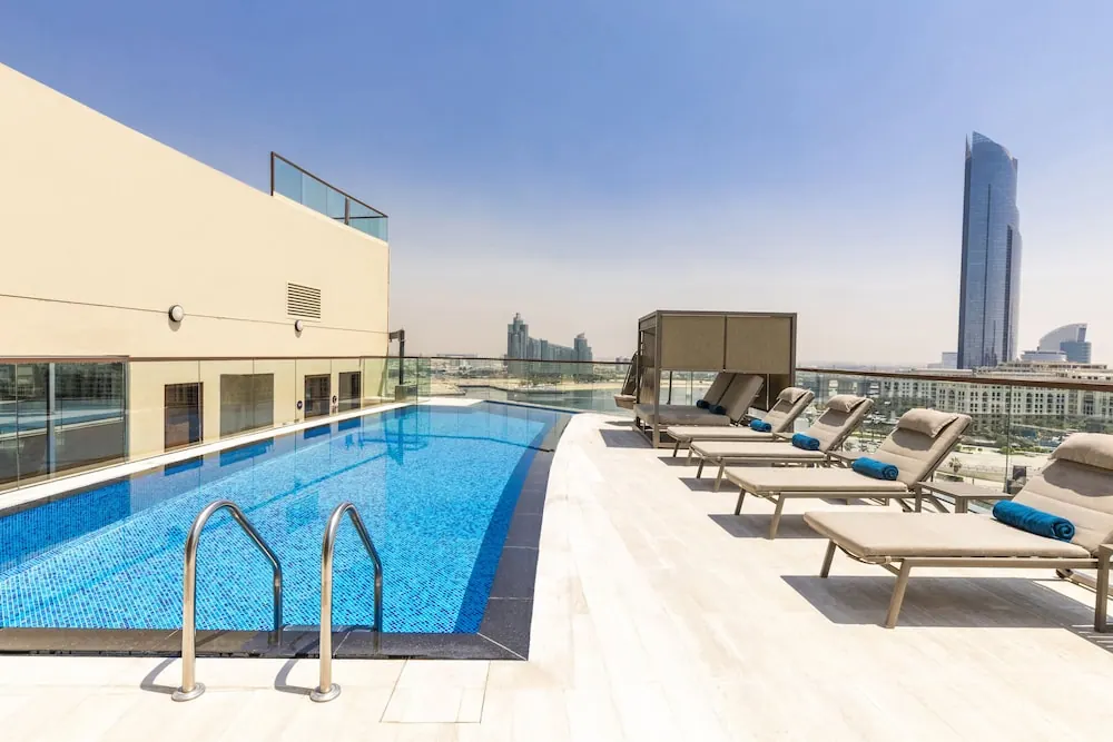 A rooftop lap pool with sun loungers and blue towels, with a city skyline and a tall glass tower in the distance.