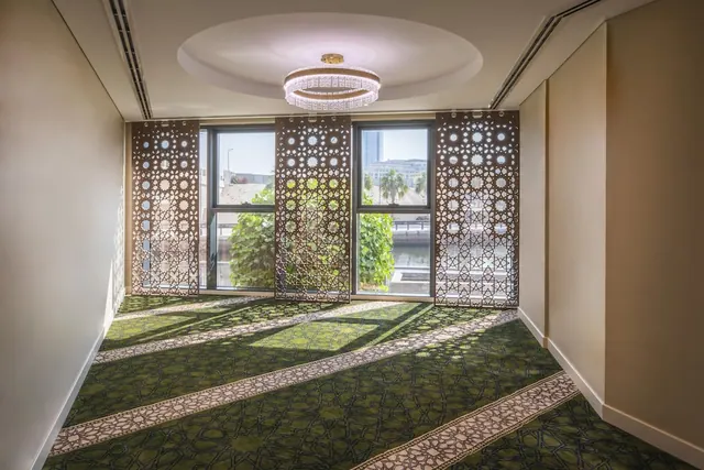 An empty room with a patterned green carpet, a ringed chandelier, decorative lattice screens and two windows looking onto greenery, water and buildings.