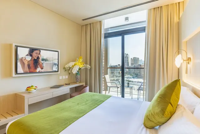 A hotel bedroom with a double bed, a wall-mounted television, a writing desk, and a glass door opening onto a balcony with chairs and city buildings beyond.