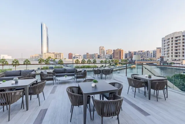 An outdoor rooftop terrace with dark tables and rope-backed chairs, lounge sofas, and a glass railing overlooking a canal toward a tall tower and city buildings.