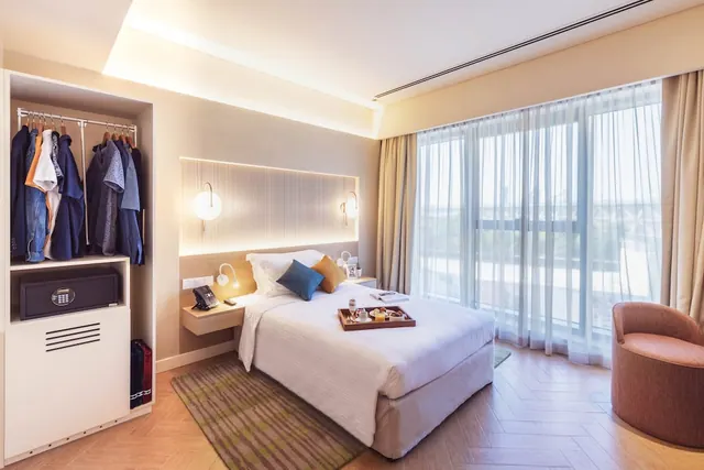 A hotel bedroom with a double bed, an open wardrobe with a safe, an armchair and floor-to-ceiling windows with sheer curtains.