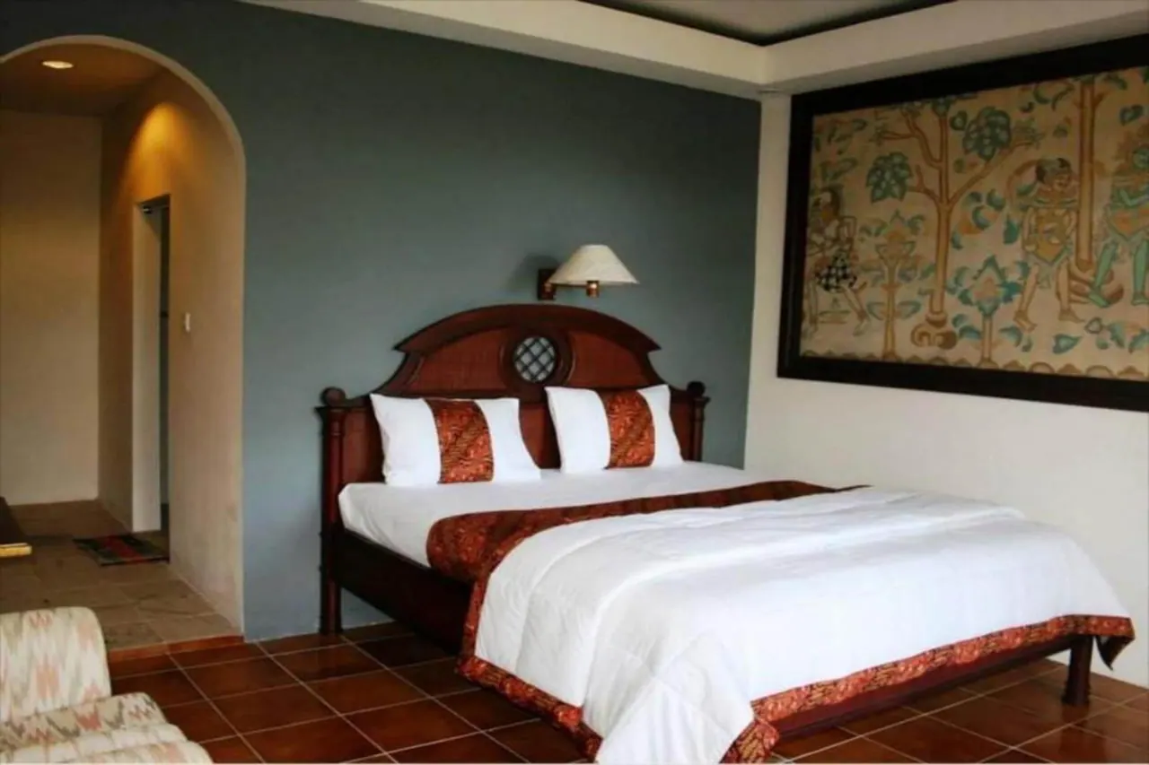 A bedroom with a double bed with white bedding and a red patterned runner, a wall lamp above the headboard, a large framed painting on the right, and a terracotta tiled floor, with an arched doorway to a hallway on the left.
