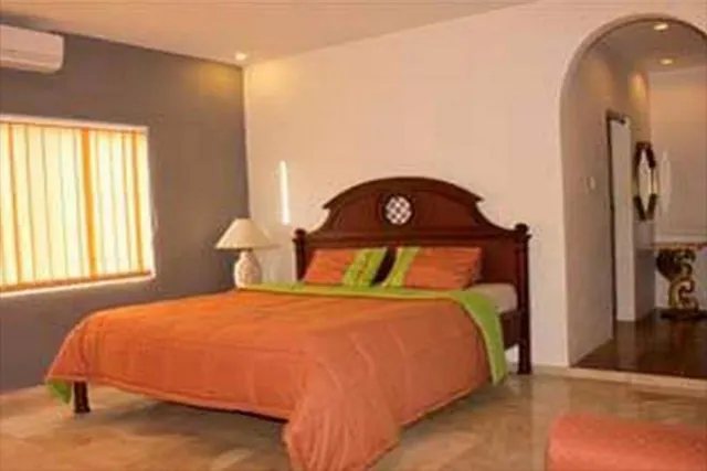 A bedroom with an orange bedspread and green trim on a dark wood bed, a table lamp on a nightstand, a window with vertical blinds, and a wall-mounted air conditioner.