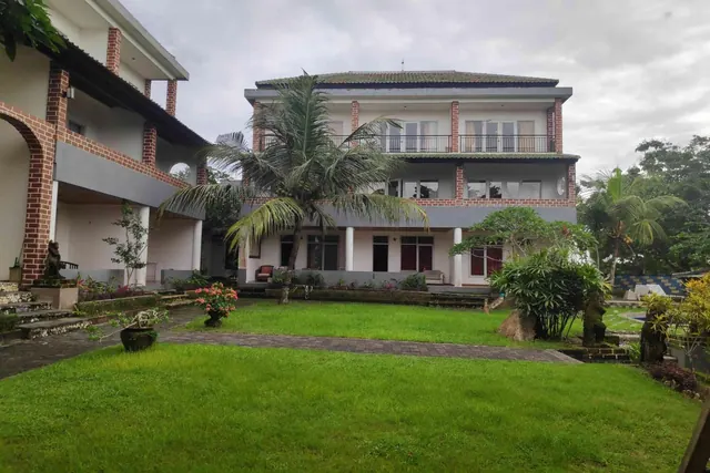 A two-storey white building with brick accents and an upper balcony stands behind a grassy lawn and a palm tree under an overcast sky.