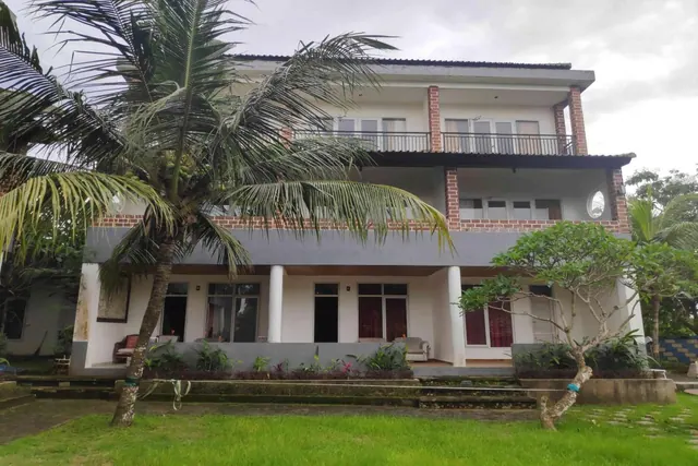 Exterior of a two-storey white building with a grey lower band, an upper balcony with railings, a palm tree in front and a grass lawn.