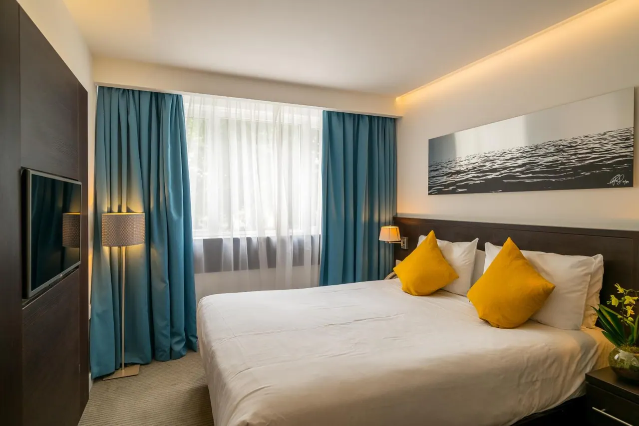 A hotel bedroom with a double bed, yellow cushions, teal curtains, a wall-mounted television and a black and white water photograph above the bed.